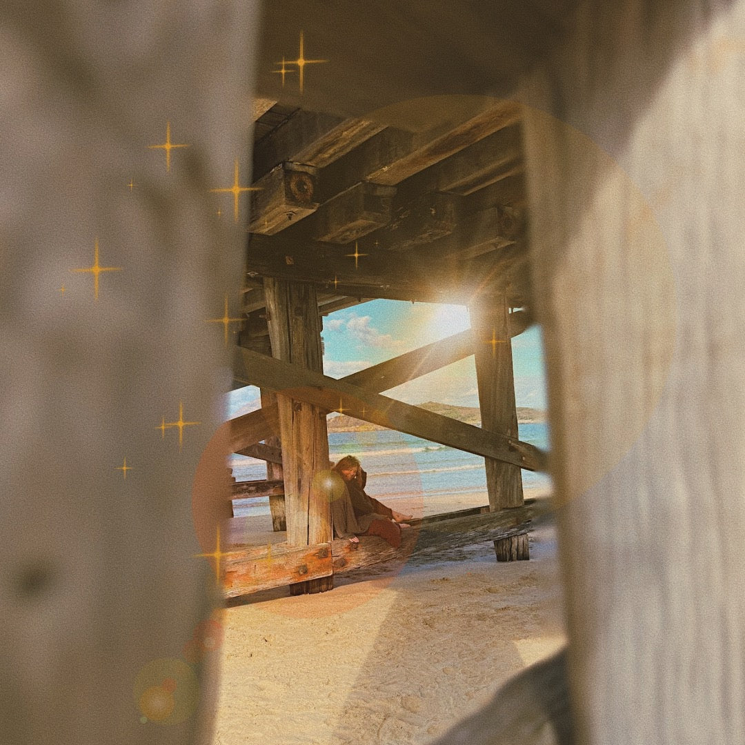 Earthy Narelle sitting under a wooden pier on a beach with sunlight filtering through.